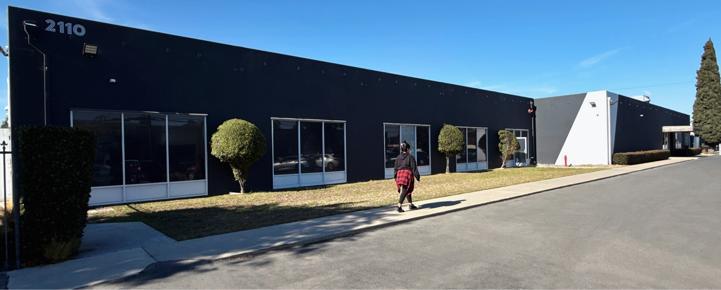A long, single-story commercial building with a black exterior, large glass windows, and the number 2110 on the upper left. A person is walking on the sidewalk in front of the building under a clear blue sky.